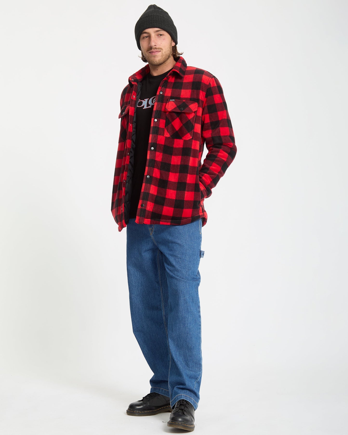 A person stands against a white background wearing a black beanie, Volcom Men's Bowered Fleece Long Sleeve in red and black plaid over a black T-shirt, loose blue jeans, and black shoes.