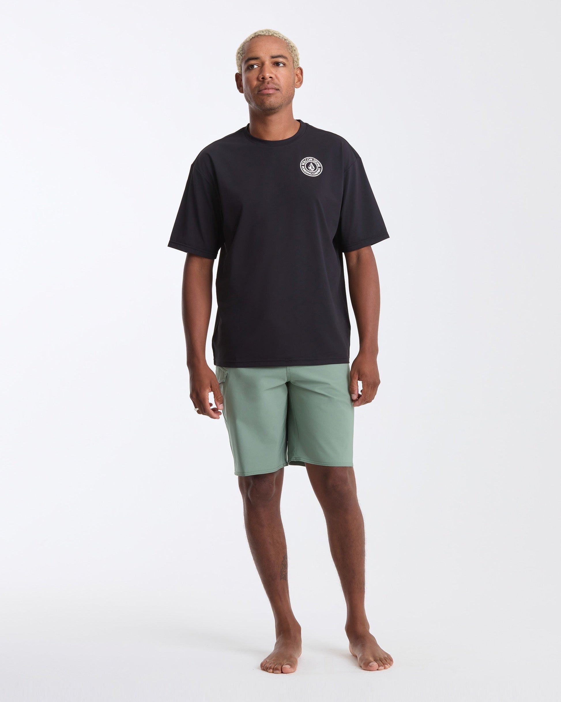 A man with short blonde hair stands barefoot against a white background, wearing Volcom's Men's Drain You Short Sleeve Tee in black with a small white chest logo and light green shorts—ideal for aquatic adventures.