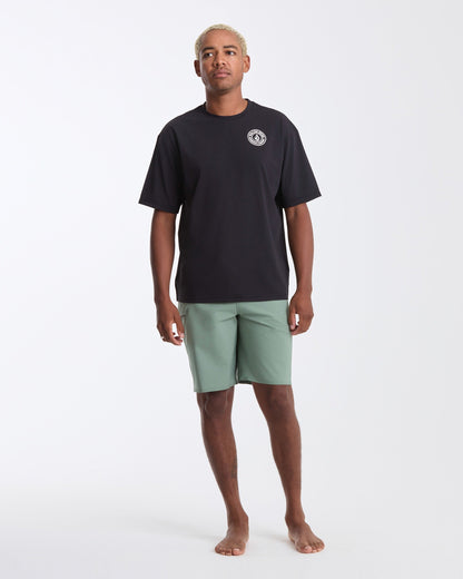A man with short blonde hair stands barefoot against a white background, wearing Volcom's Men's Drain You Short Sleeve Tee in black with a small white chest logo and light green shorts—ideal for aquatic adventures.