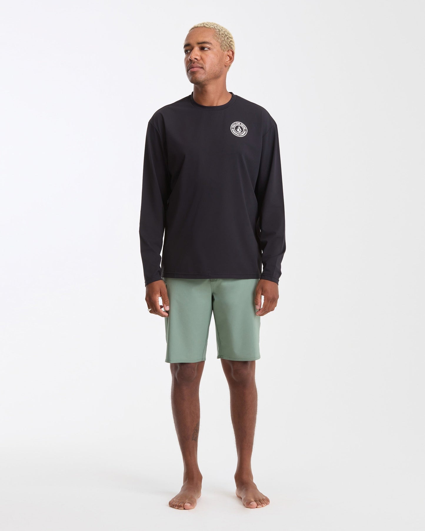 A man with short blond hair stands barefoot against a plain white background, wearing the Volcom Men's Drain You Long Sleeve black rashguard with UV protection and a small white chest logo, paired with light green shorts.