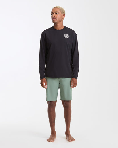 A man with short blond hair stands barefoot against a plain white background, wearing the Volcom Men's Drain You Long Sleeve black rashguard with UV protection and a small white chest logo, paired with light green shorts.