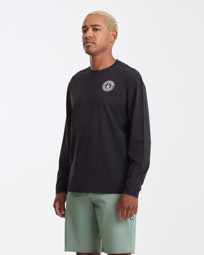 A man with short blond hair wears the Volcom Men's Drain You Long Sleeve rashguard in a boxy fit, featuring a small white circular chest logo and light green shorts, standing against a plain white background.