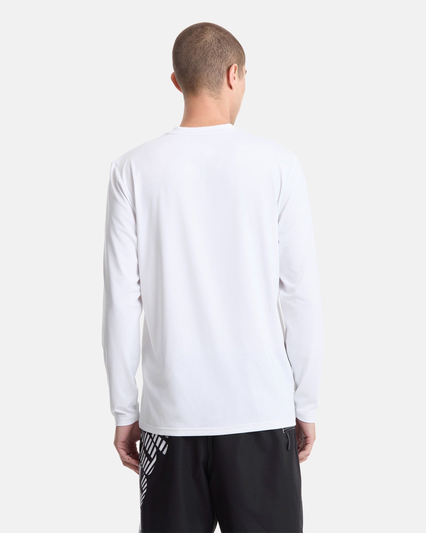 A person with short hair wears a Volcom Men's Modstone Loose Long Sleeve rashguard in white and black graphic shorts, standing with their back to a plain light background.