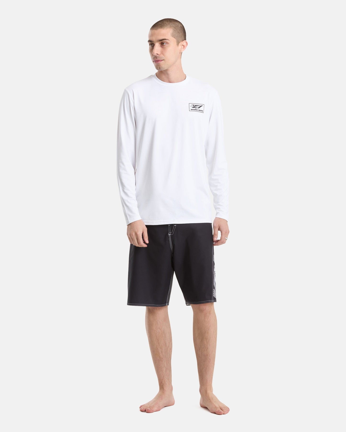 A man stands barefoot against a light background, wearing the Volcom Men's Modstone Loose Long Sleeve rashguard with a small chest logo and black knee-length shorts with white stitching, perfect for UV protection.
