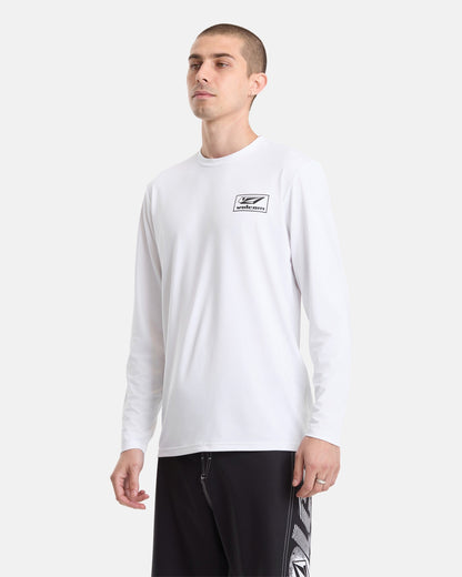 A man stands against a plain light background wearing a white Volcom Men's Modstone Loose Long Sleeve shirt with UV protection, featuring a small black chest logo, paired with black shorts with a white pattern.
