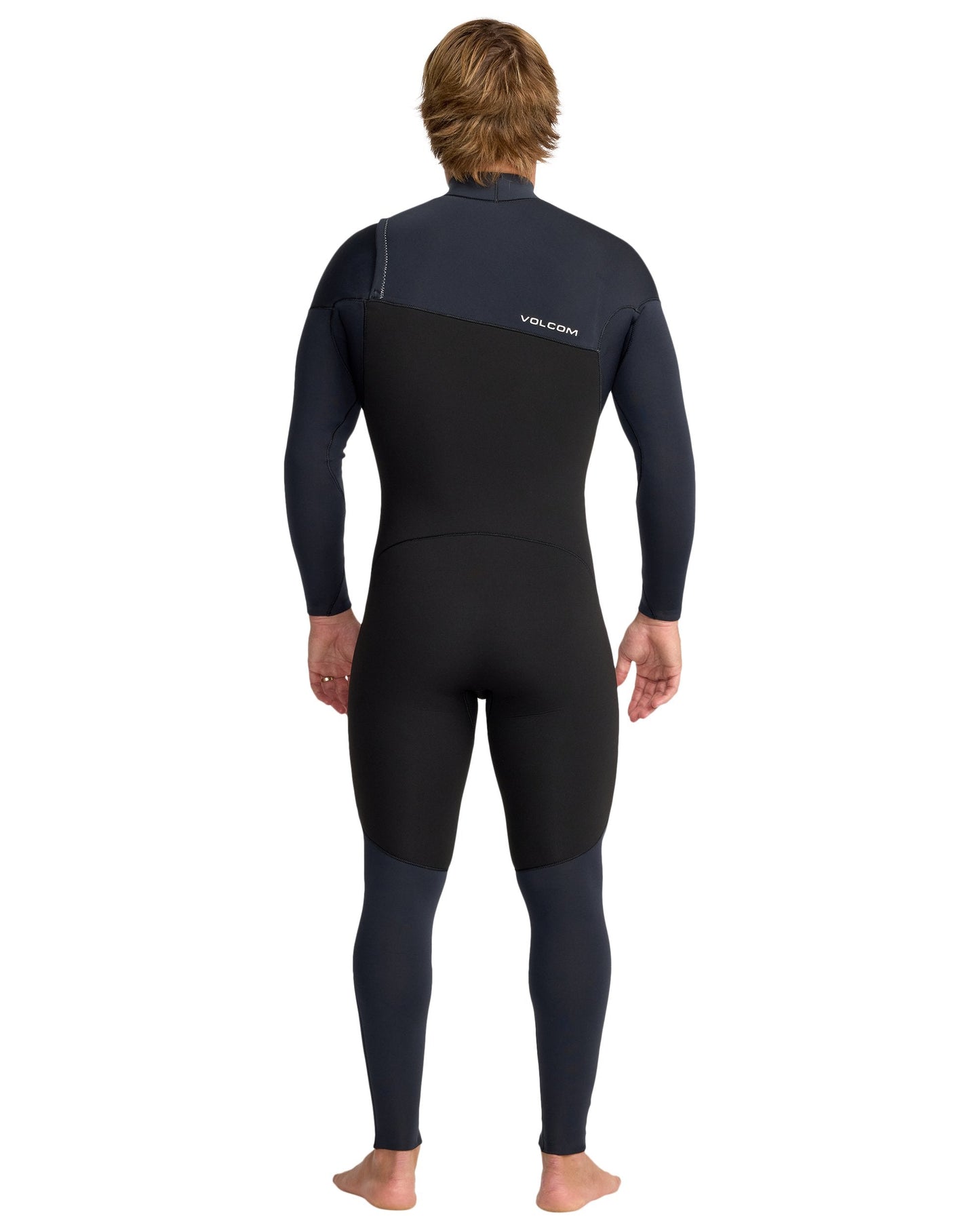 A person with light brown hair, barefoot and facing away, wears the Volcom Men's 4/3 Modulator Chest Zip Full Wetsuit in black and dark navy, featuring long sleeves and the Volcom logo on the upper back.