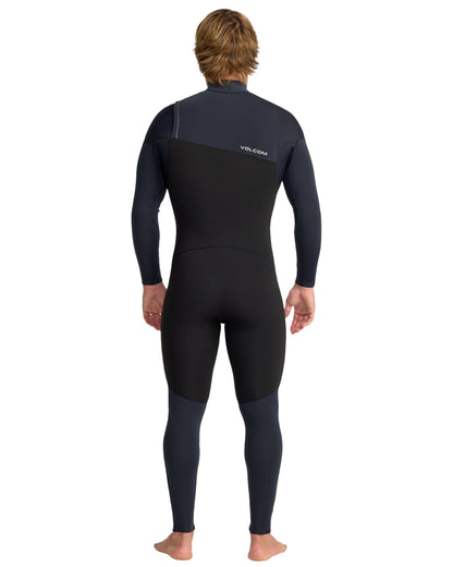 A person with light brown hair, barefoot and facing away, wears the Volcom Men's 4/3 Modulator Chest Zip Full Wetsuit in black and dark navy, featuring long sleeves and the Volcom logo on the upper back.