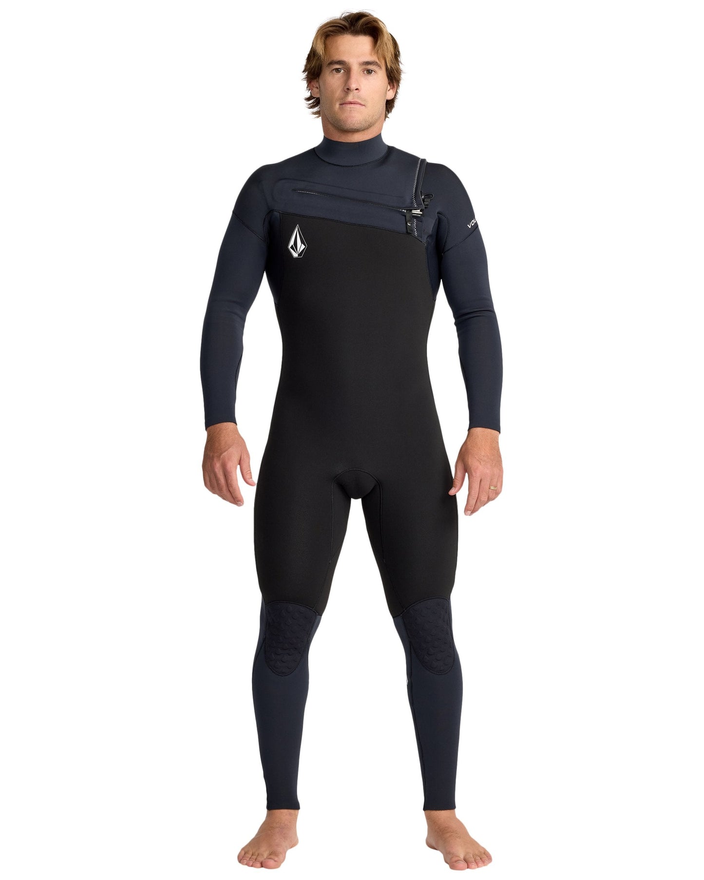 A man with light brown hair stands barefoot, facing forward, in the Volcom Men's 4/3 Modulator Chest Zip Full Wetsuit—black and dark gray with long sleeves and a small logo on the chest. His arms are relaxed at his sides.