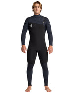 A man with light brown hair stands barefoot, facing forward, in the Volcom Men's 4/3 Modulator Chest Zip Full Wetsuit—black and dark gray with long sleeves and a small logo on the chest. His arms are relaxed at his sides.