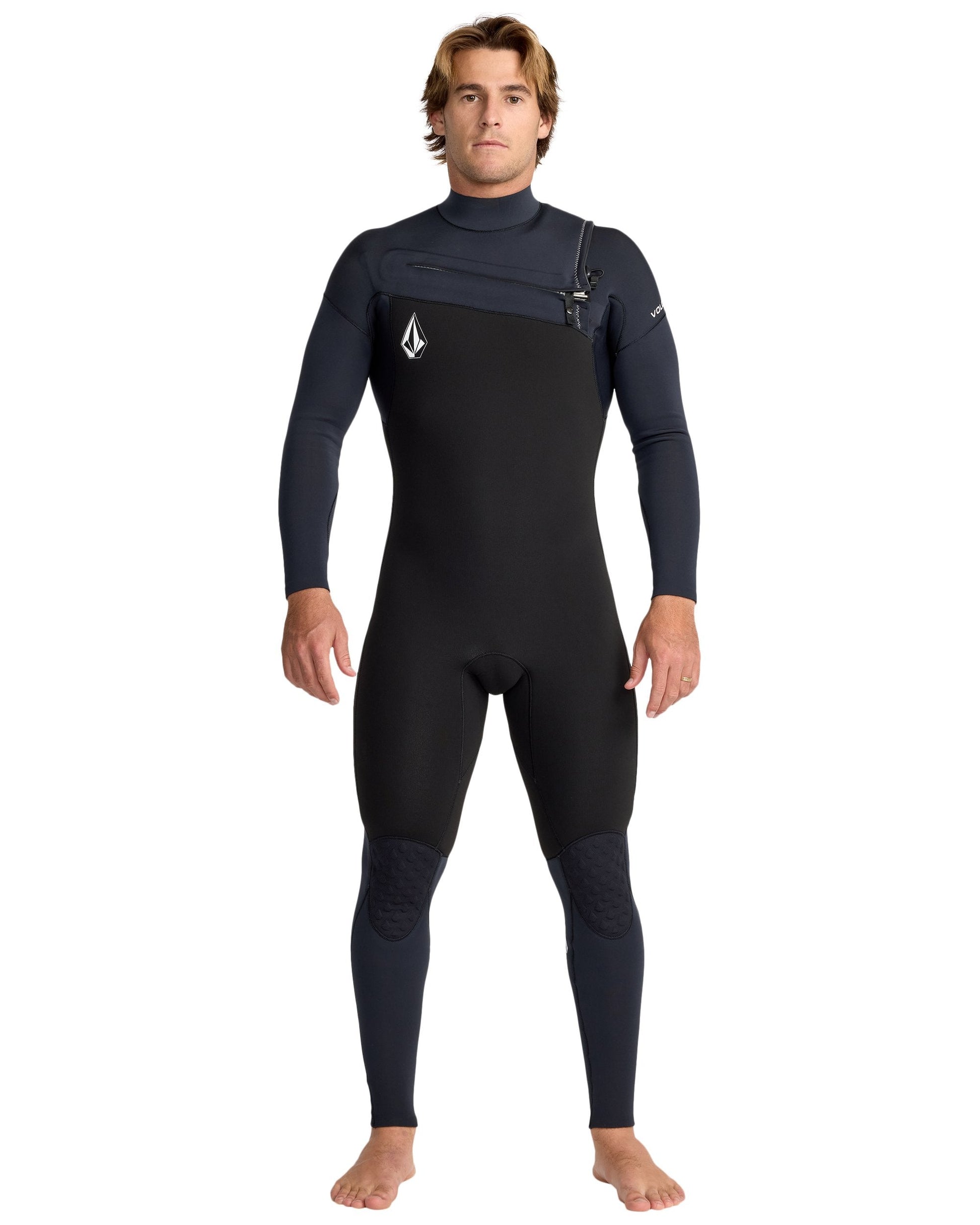 A man with light brown hair stands barefoot, facing forward, in the Volcom Men's 4/3 Modulator Chest Zip Full Wetsuit—black and dark gray with long sleeves and a small logo on the chest. His arms are relaxed at his sides.