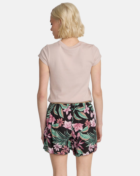A woman with short blonde hair is seen from behind wearing a light pink short-sleeve shirt and Volcom Women's Boss Beach Shorts, which feature a black base with a vibrant pink and green floral design.