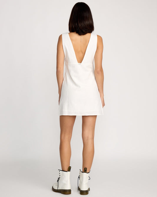 A woman with straight, shoulder-length dark hair is seen from behind in a plain setting, wearing the Volcom Women's Money Mama Dress—a sleeveless short white dress with a deep V-cut back—paired with white ankle-high lace-up boots.