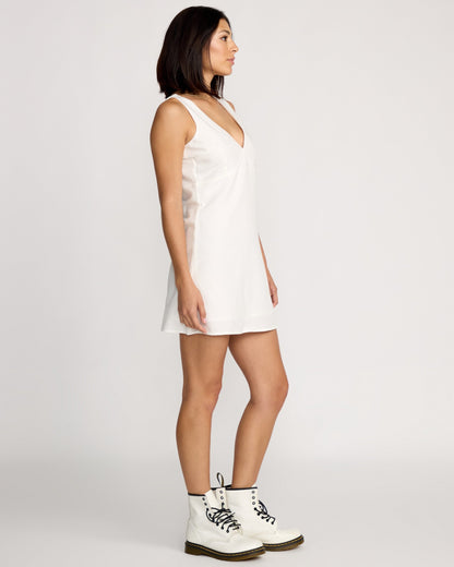 A woman with straight shoulder-length dark hair stands in profile against a plain background, wearing the Volcom Women's Money Mama Dress, a sleeveless white V-neck mini dress, paired with white lace-up ankle boots.