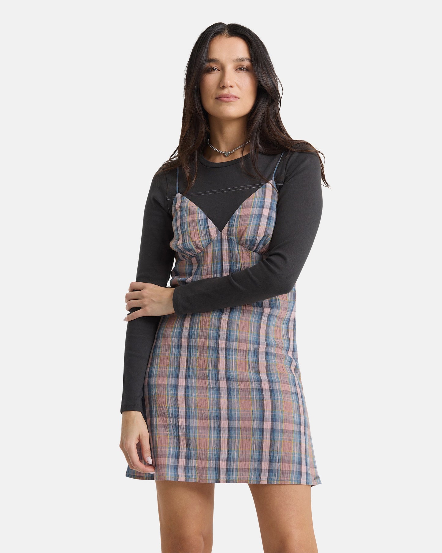 A woman with long dark hair faces forward, wearing the Volcom Women's Sand Burn Dress over a long-sleeve black shirt. She poses with a neutral expression and one arm across her body against a plain white background.