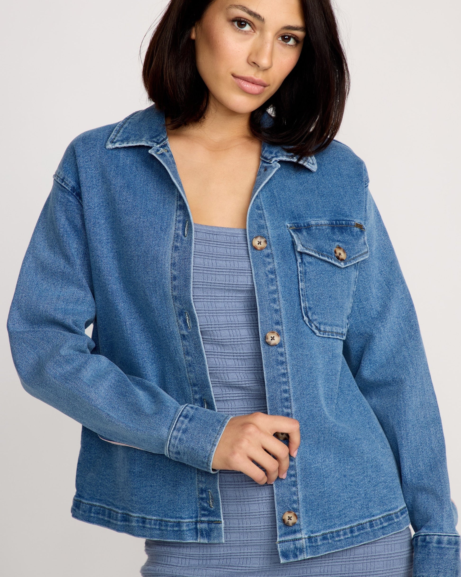 A woman with shoulder-length dark hair models the Volcom Women's Stoned BF Shacket over a textured, sleeveless light blue dress, standing against a plain, light-colored background.