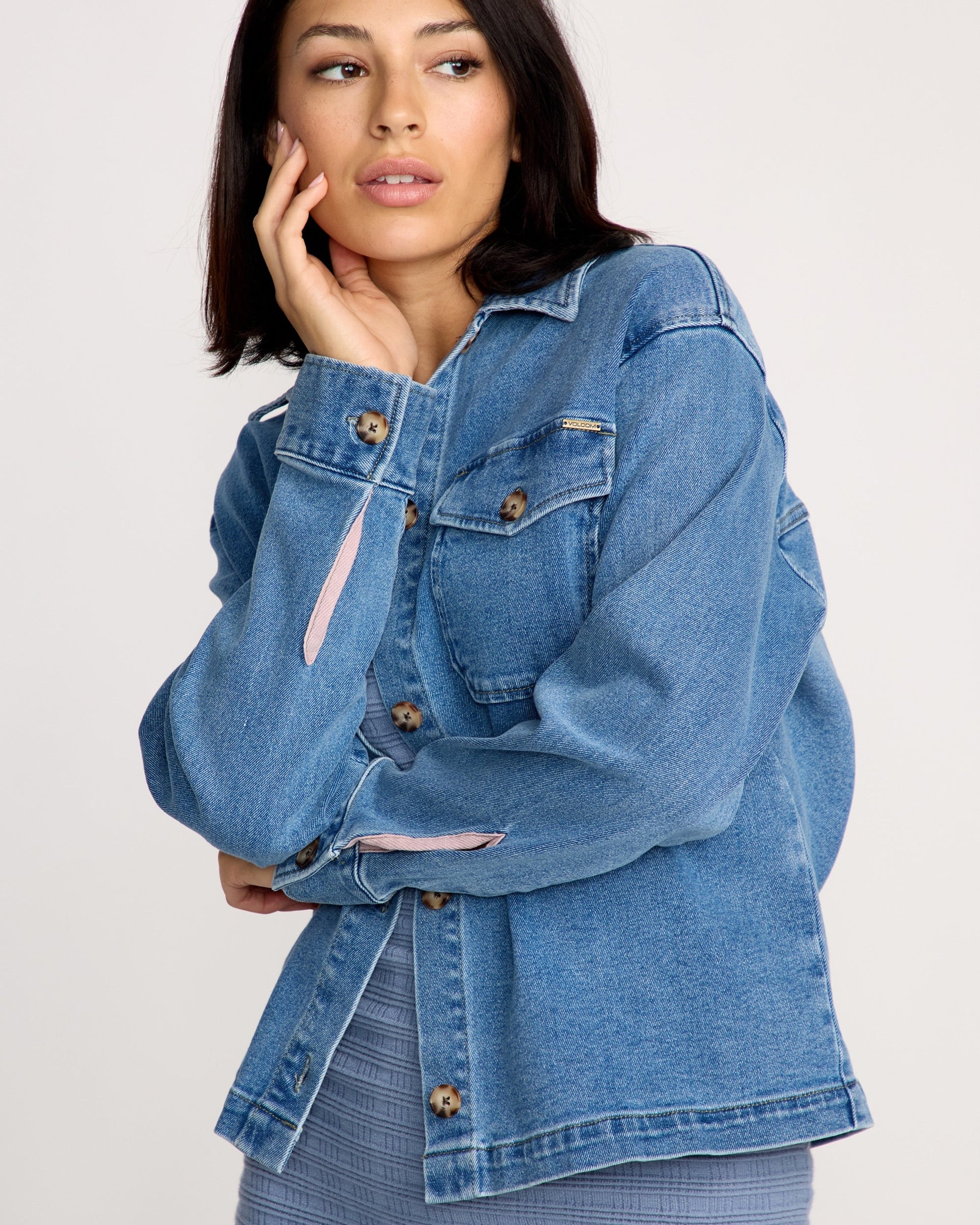 A woman with straight dark hair wears the Volcom Women's Stoned BF Shacket over a ribbed gray top, gazing to the side with her hand on her cheek against a simple, light-colored background.