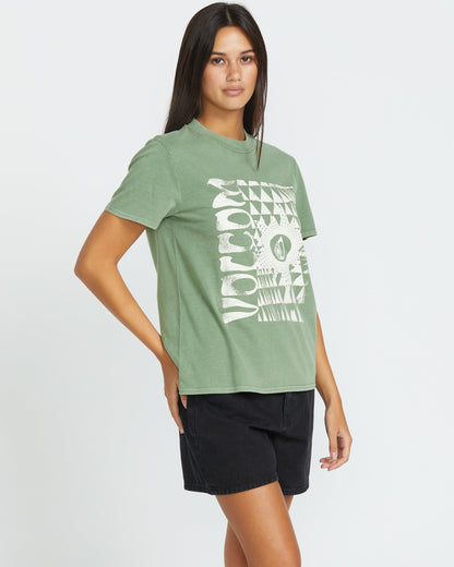 A woman with long dark hair poses against a white background wearing the Women's Lock It Up Tee by Volcom—an organic cotton green t-shirt with a white geometric graphic—and black shorts, standing with one hand on her hip.