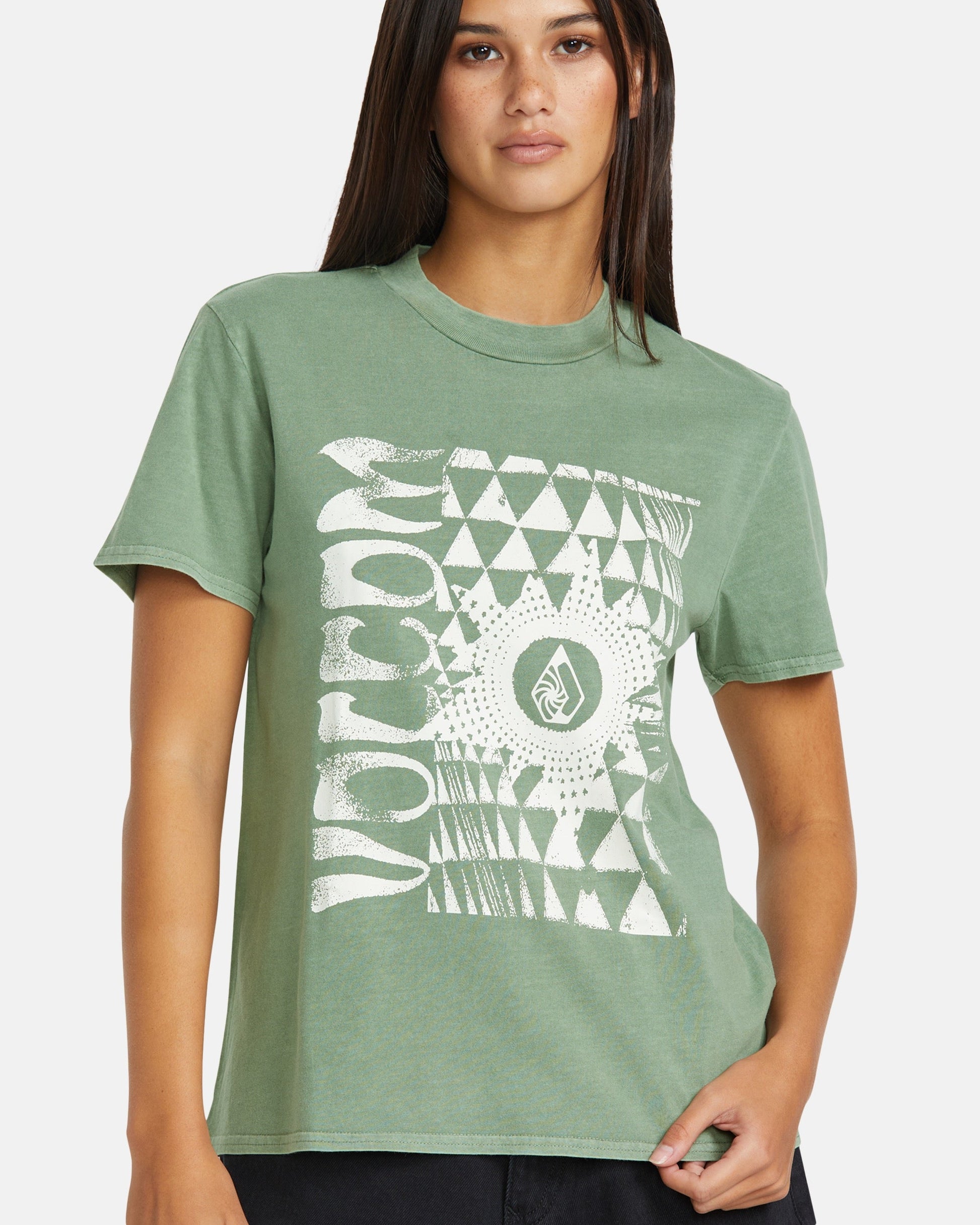 A woman models the Volcom Women’s Lock It Up Tee, a green relaxed-fit graphic tee made from organic cotton with a white abstract geometric design and "VOLCOM" on the front, standing against a plain background.