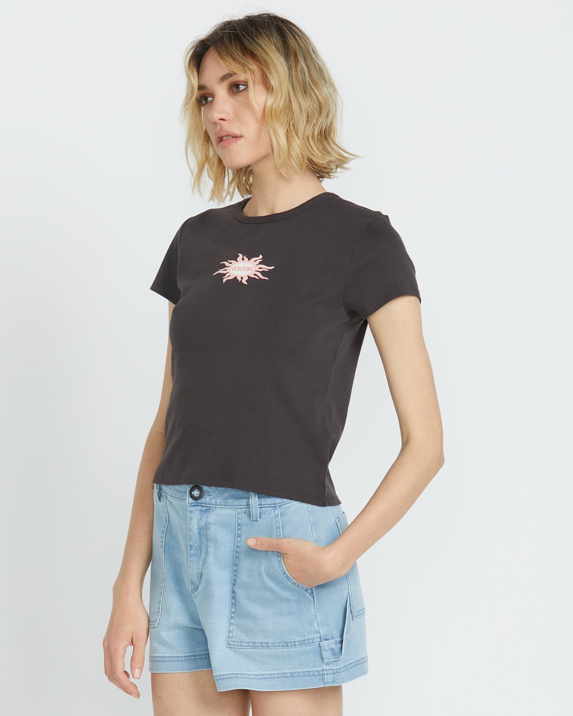 A woman with wavy blonde hair wears the Volcom Women's Have A Clue Tee, featuring a pink chest graphic, paired with light blue denim shorts. She stands against a plain white background, her left hand in her pocket.