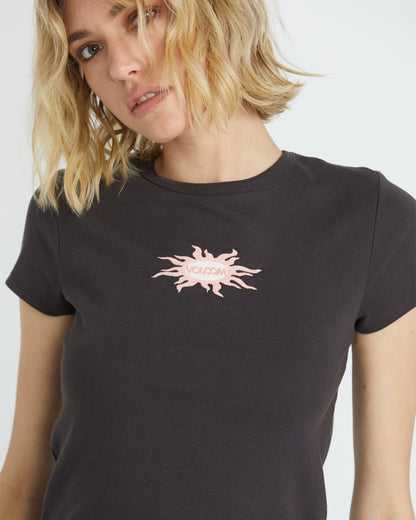 A woman with blonde hair models the Volcom Women's Have A Clue Tee, a black casual shirt with a pink abstract sunburst and "Volcom" on the front, looking to the side with a neutral expression.