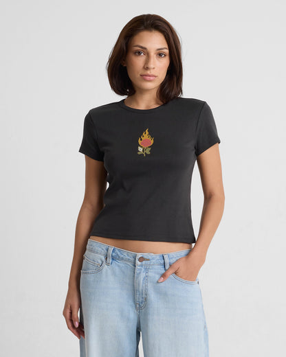 A woman with straight brown hair wears the Volcom Women's Have A Clue Tee, a fitted black t-shirt with a small flaming torch graphic on the chest, paired with light blue jeans. She stands against a plain white background with a neutral expression.
