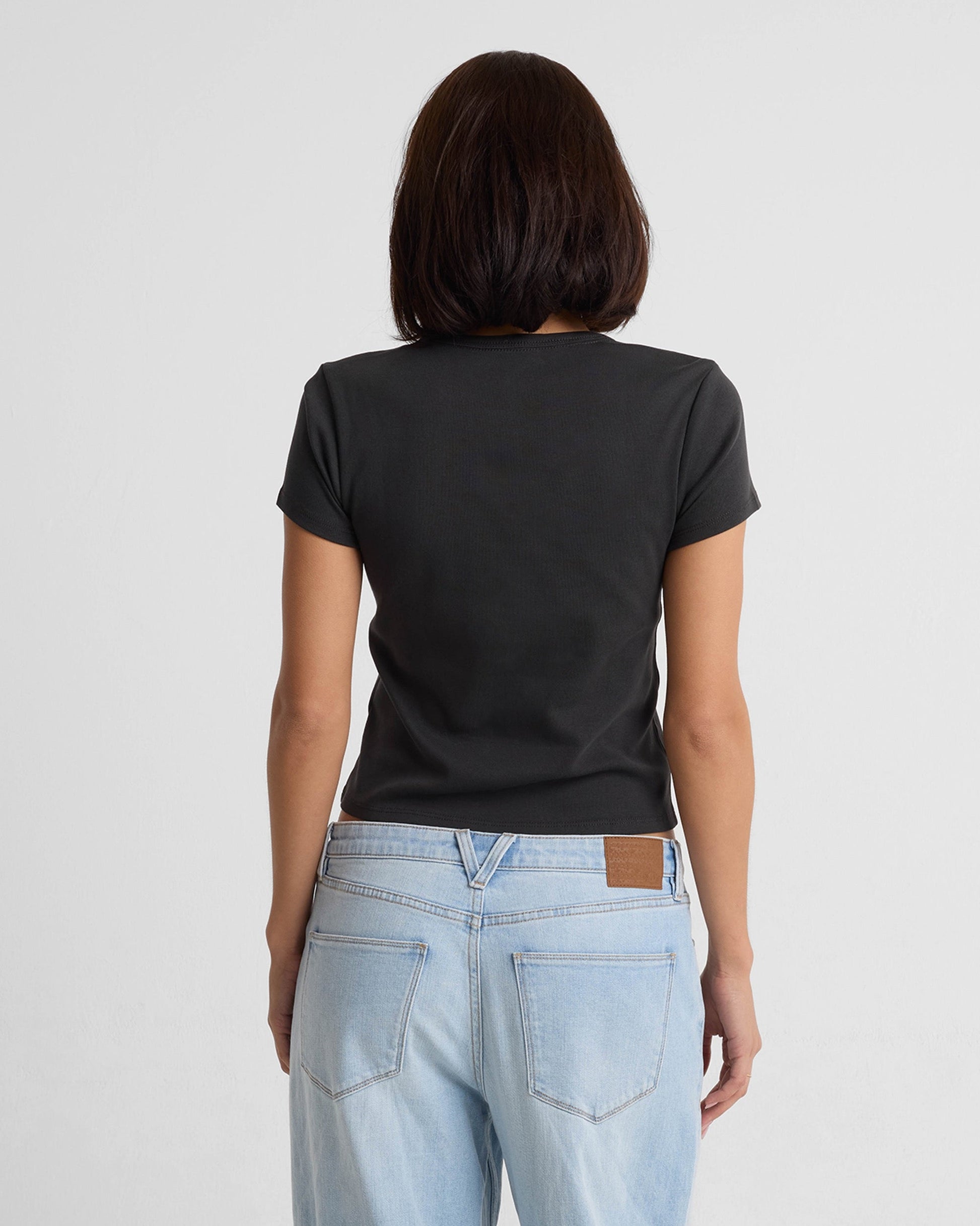 A woman with shoulder-length dark hair, seen from behind, wears the Volcom Women's Have A Clue Tee in black and light blue jeans against a plain white background.