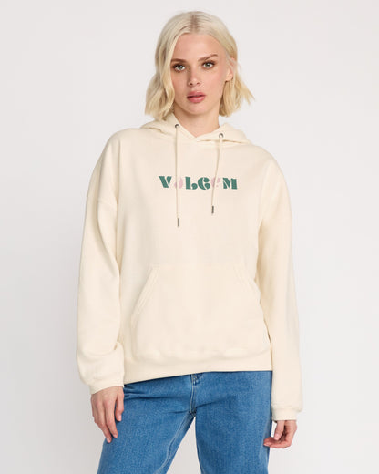 A woman with short blonde hair models the Volcom Women's Stone Art Up Hoodie, a cream-colored oversized style with green and pink VOLCOM logo, paired with blue jeans against a white background—ideal for relaxed streetwear looks.