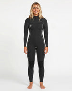 A person stands facing forward, smiling and barefoot on a plain white background, wearing the Volcom Women's 4/3MM Modulator Chest Zip Hooded Full Wetsuit with white seams and knee pads.