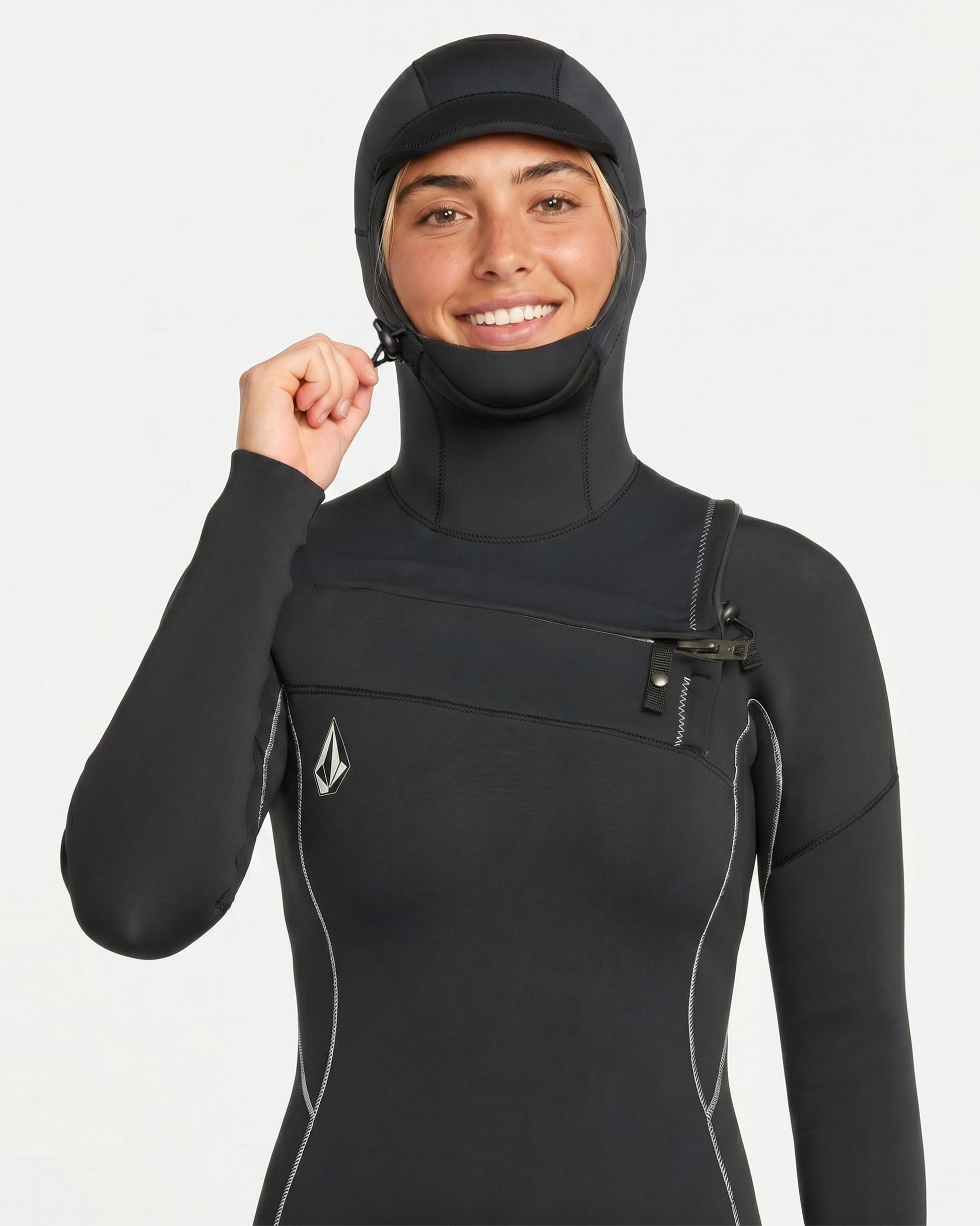 A person in the Volcom Women's 4/3MM Modulator Chest Zip Hooded Full Wetsuit, featuring white stitching and a small chest logo, smiles at the camera while pulling on the hood’s zip against a plain white background.