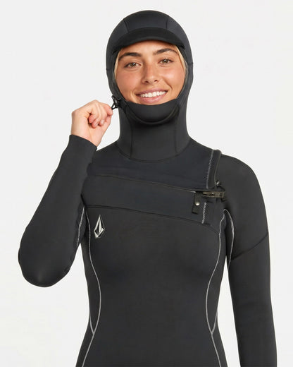 A person in the Volcom Women's 4/3MM Modulator Chest Zip Hooded Full Wetsuit, featuring white stitching and a small chest logo, smiles at the camera while pulling on the hood’s zip against a plain white background.