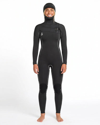 A person stands barefoot and smiling against a plain white background, wearing the Volcom Women's 4/3MM Modulator Chest Zip Hooded Full Wetsuit with white seams.