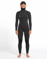 A person stands barefoot and smiling against a plain white background, wearing the Volcom Women's 4/3MM Modulator Chest Zip Hooded Full Wetsuit with white seams.