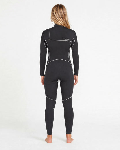 A person with long blonde hair stands barefoot facing away in a Volcom Women's 4/3MM Modulator Chest Zip Hooded Full Wetsuit, featuring white seam lines and "VOLCOM" on the upper back, against a plain white background.