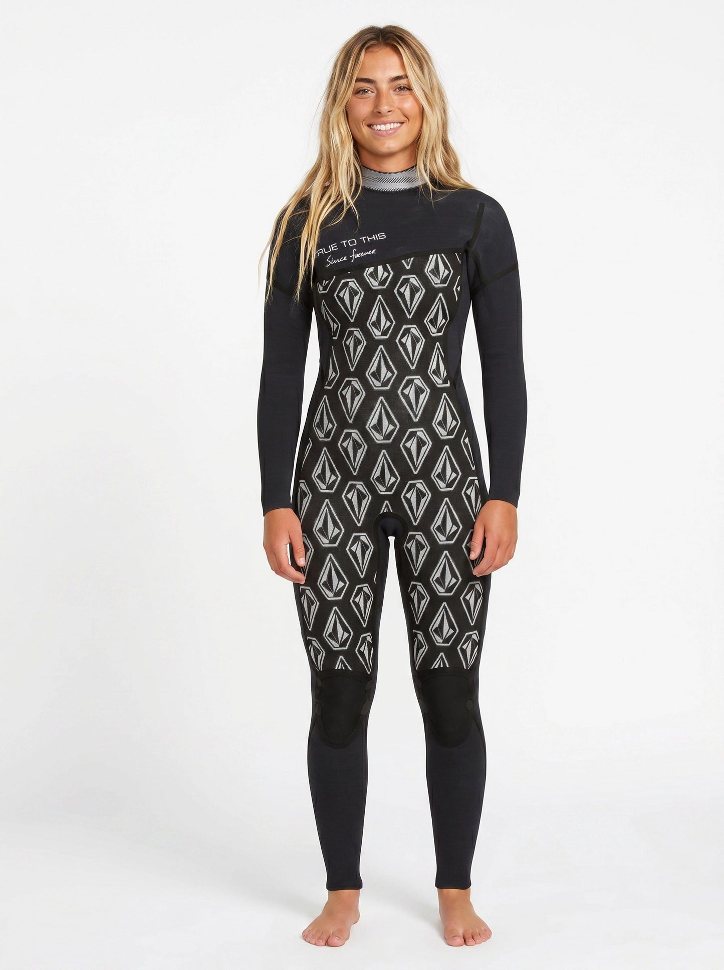 A smiling woman with long blonde hair stands barefoot against a white background, wearing the Volcom Women's 3/2MM Modulator Chest Zip Full Wetsuit featuring white geometric patterns.