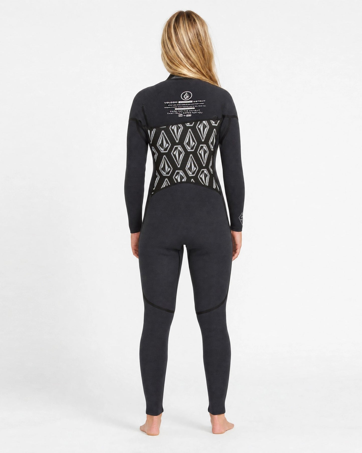 A woman with long blonde hair stands barefoot in a Volcom Women's 3/2MM Modulator Chest Zip Full Wetsuit, black with a geometric pattern and white back text, against a plain white background.