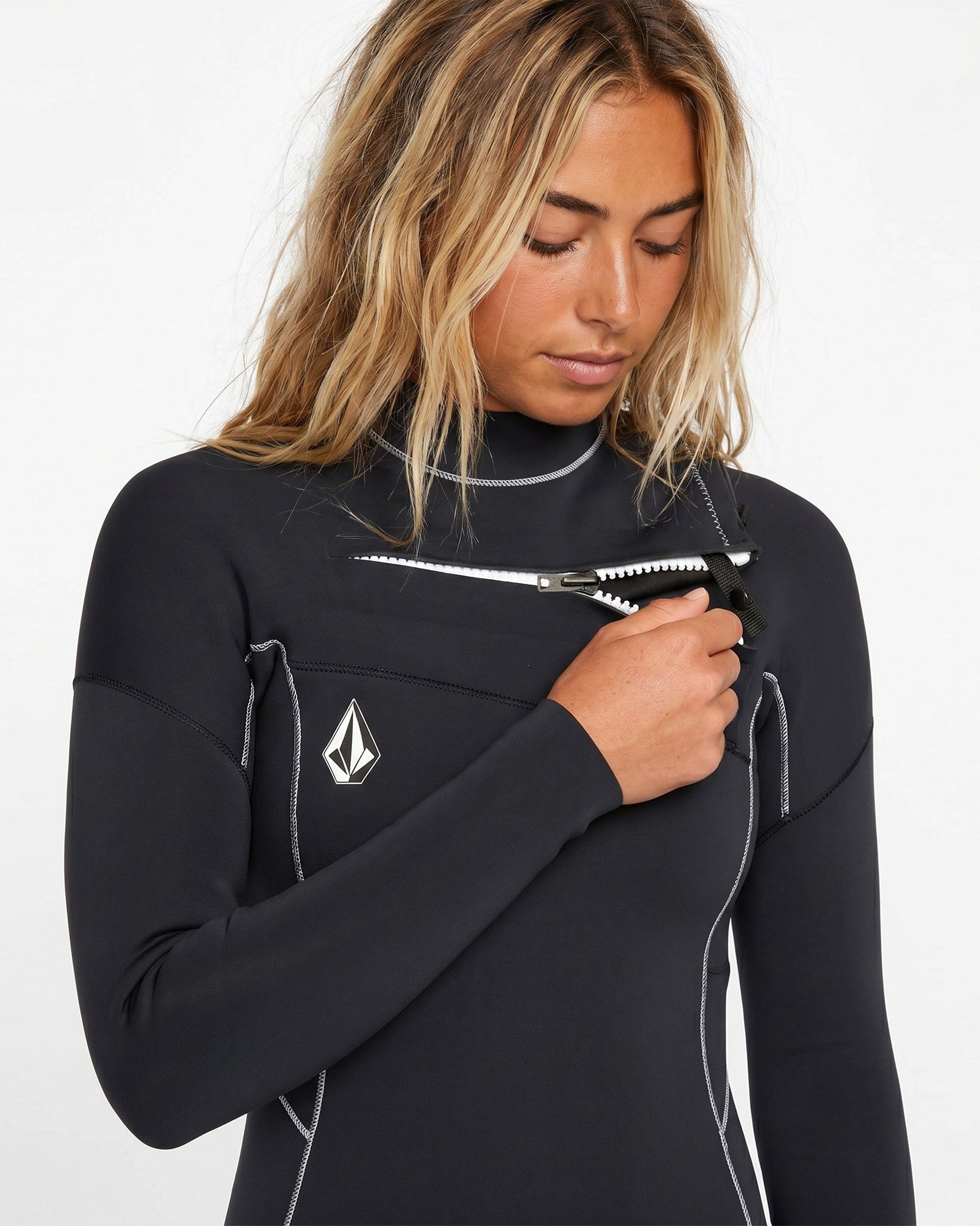 A person in a black Volcom Women's 3/2MM Modulator Chest Zip Full Wetsuit with white stitching and a geometric chest logo holds a serrated knife pointed downward, eyes lowered, with long blonde hair worn loose.