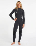 A woman with long blonde hair smiles barefoot in a Volcom Women's 3/2MM Modulator Chest Zip Full Wetsuit with white seams and reinforced knee patches, standing against a plain white background.