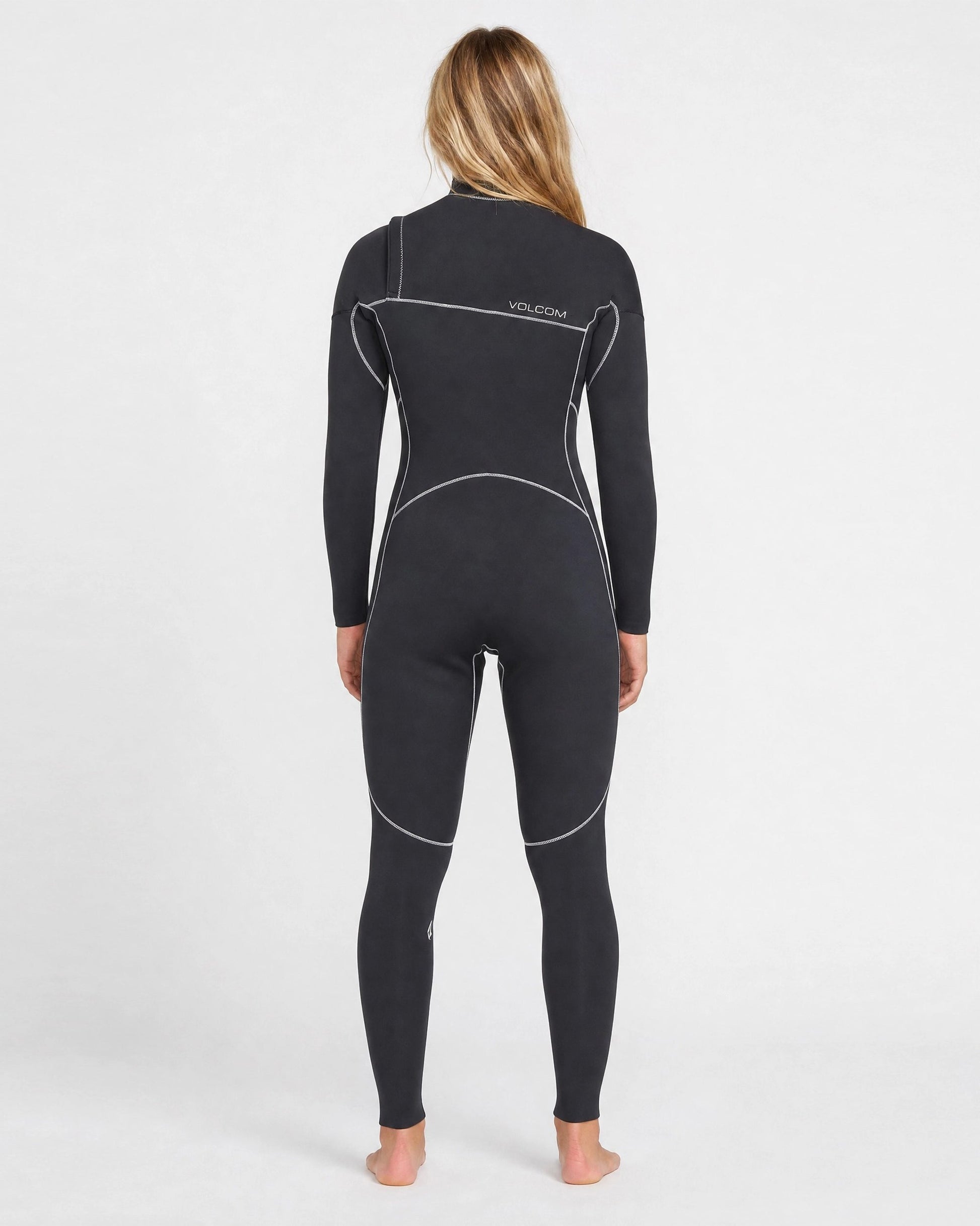 A person with long blonde hair stands barefoot, facing away, wearing a black Volcom Women's 3/2MM Modulator Chest Zip Full Wetsuit with white seams and “Volcom” on the back, against a plain white background.