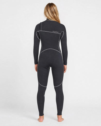 A person with long blonde hair stands barefoot, facing away, wearing a black Volcom Women's 3/2MM Modulator Chest Zip Full Wetsuit with white seams and “Volcom” on the back, against a plain white background.
