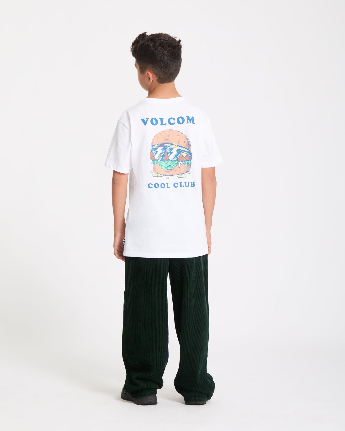 A boy faces away, wearing the Kids' Burgstone Pocket Tee—an organic cotton white T-shirt with burger and ocean art plus "VOLCOM COOL CLUB" text—paired with dark green pants and black shoes, set against a plain white background.