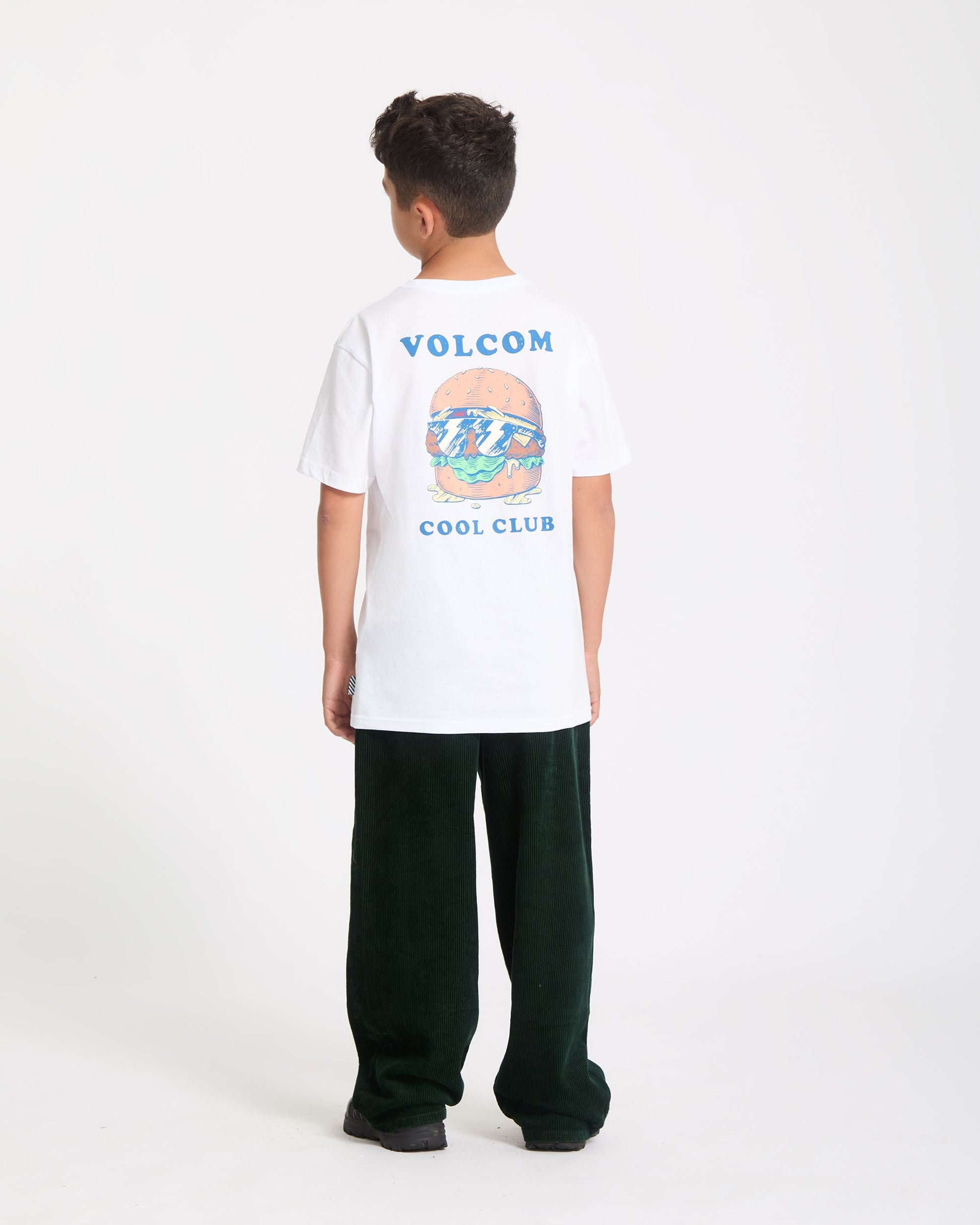 A boy faces away, wearing the Kids' Burgstone Pocket Tee—an organic cotton white T-shirt with burger and ocean art plus "VOLCOM COOL CLUB" text—paired with dark green pants and black shoes, set against a plain white background.