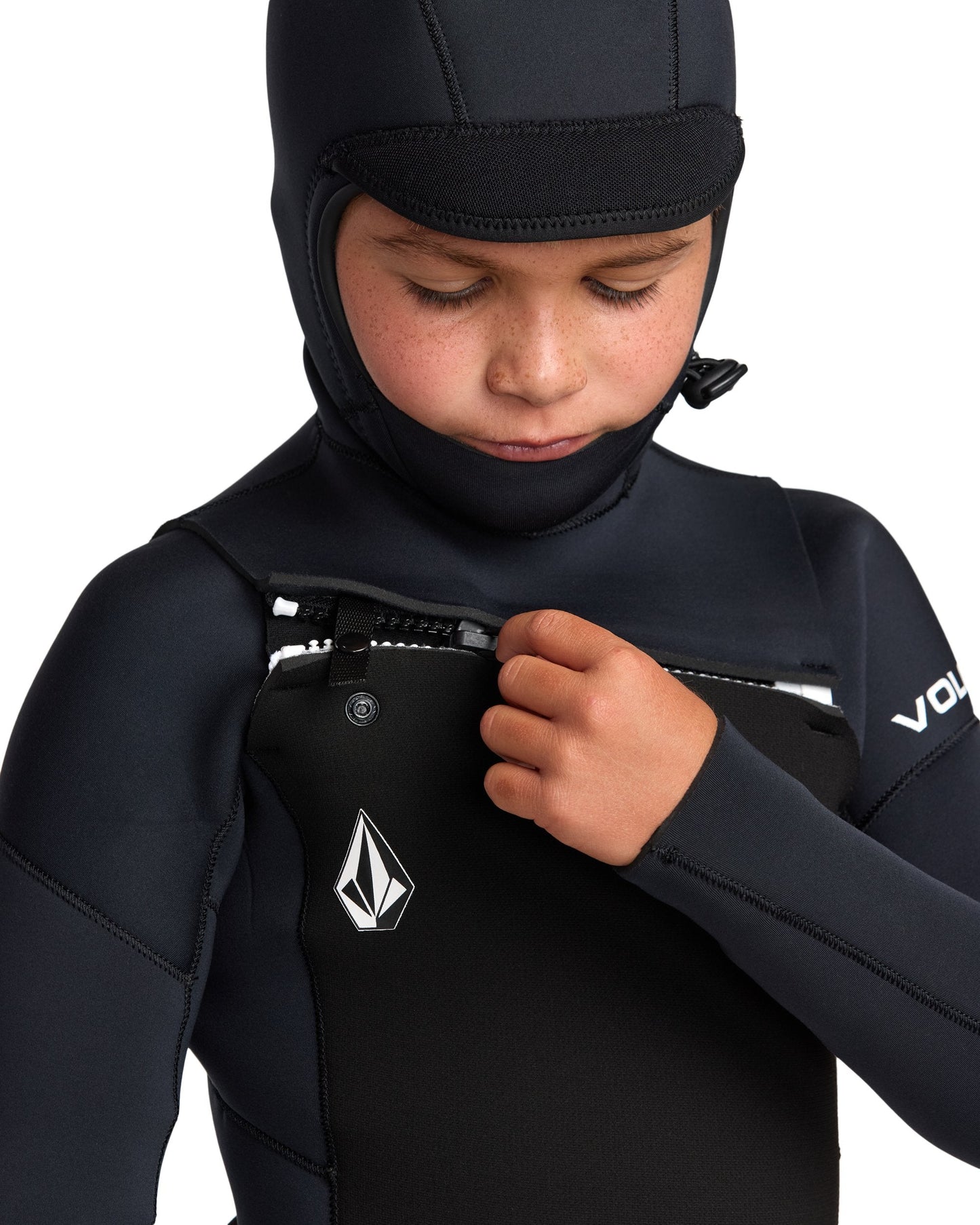 A child zips up the chest of the Volcom Kids' 5/4mm Hooded Chest Zip Long Sleeve Full Wetsuit in black, featuring a white chest logo and partial Volcom branding on the sleeve.