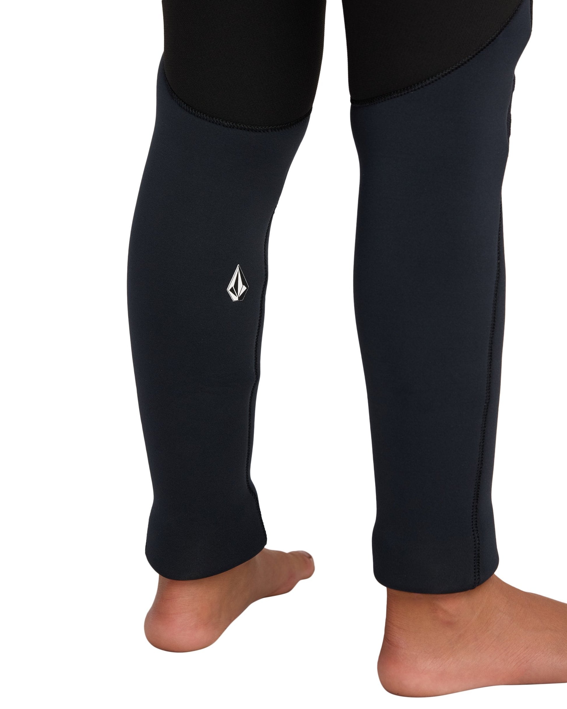 A close-up of the lower legs of a person standing barefoot, wearing the Volcom Kids' 5/4 mm Hooded Chest Zip Long Sleeve Full Wetsuit with a small white logo on one leg.