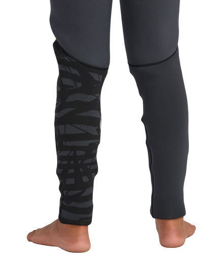 A child stands barefoot from the calves down in Volcom’s Kids' 3/2 Modulator Chest Zip Full Wetsuit, featuring dark, fitted legs—one with a black and gray abstract pattern, the other solid dark gray—ideal for young surf enthusiasts.