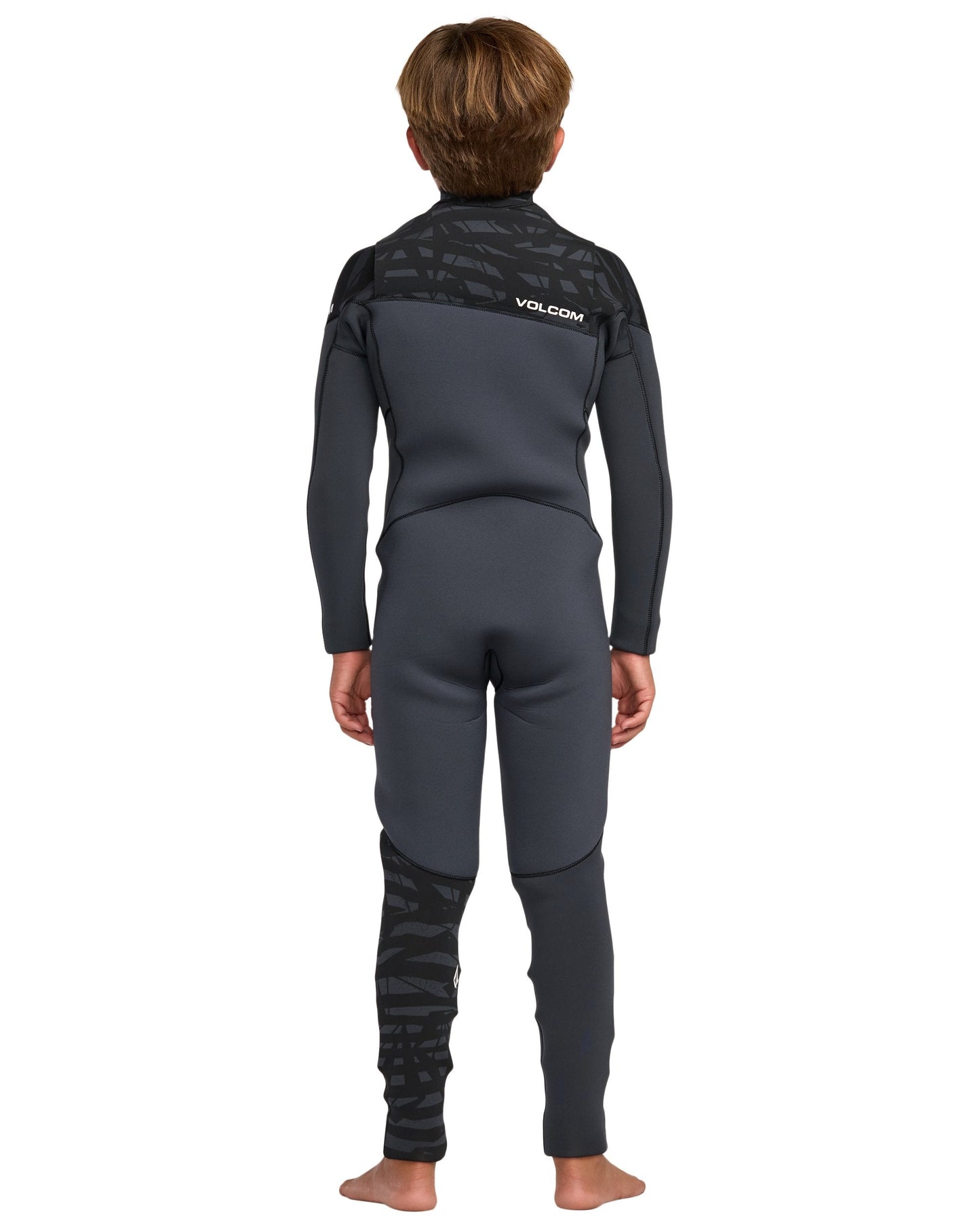 A young person with short brown hair stands barefoot, facing away, in a Volcom Kids' 3/2 Modulator Chest Zip Full Wetsuit. The wetsuit is black and dark gray with patterned details on the shoulders and lower left leg. Background is plain white.