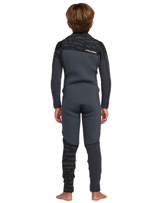 A young person with short brown hair stands barefoot, facing away, in a Volcom Kids' 3/2 Modulator Chest Zip Full Wetsuit. The wetsuit is black and dark gray with patterned details on the shoulders and lower left leg. Background is plain white.
