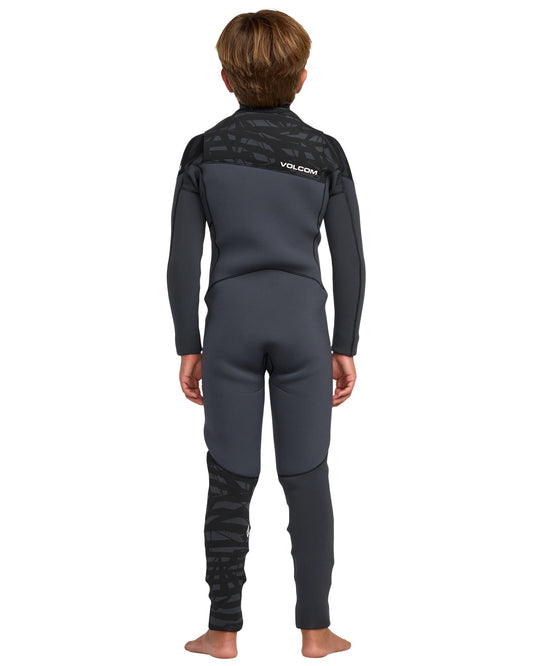 A child stands barefoot, facing away, wearing the Volcom Kids' 4/3 Modulator Chest Zip Full Wetsuit in black and dark gray with a textured pattern on the shoulders and lower left leg. The Volcom logo appears on the upper back.