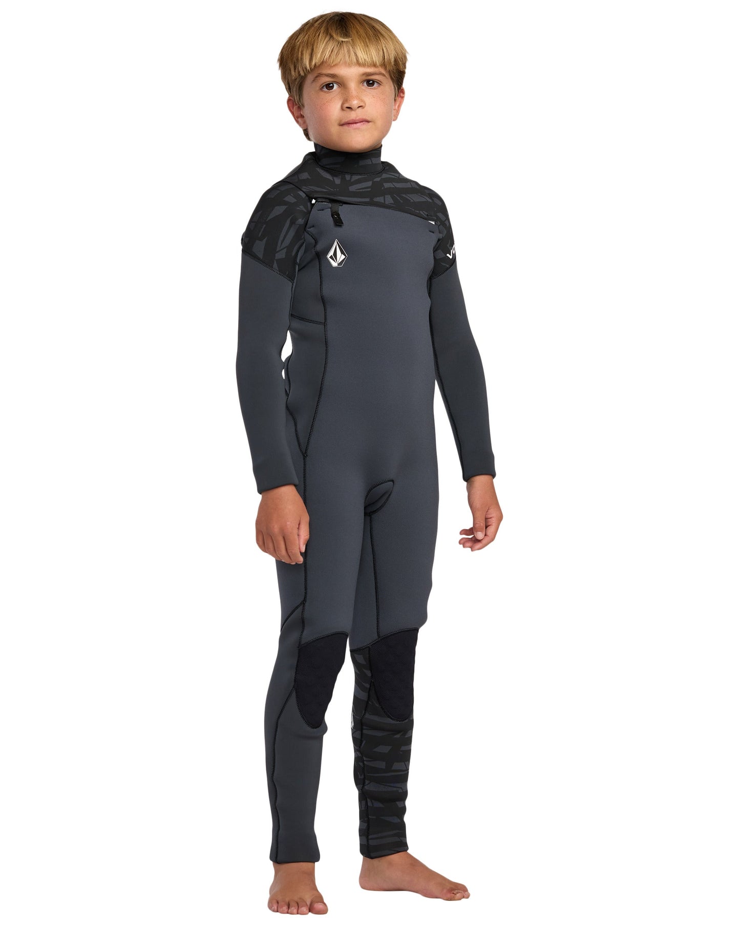 A young boy stands barefoot in the Volcom Kids' 4/3 Modulator Chest Zip Full Wetsuit with dark gray fabric, black accents, and a chest logo, posing confidently against a white background.