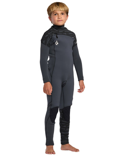 A young boy stands barefoot in the Volcom Kids' 4/3 Modulator Chest Zip Full Wetsuit with dark gray fabric, black accents, and a chest logo, posing confidently against a white background.