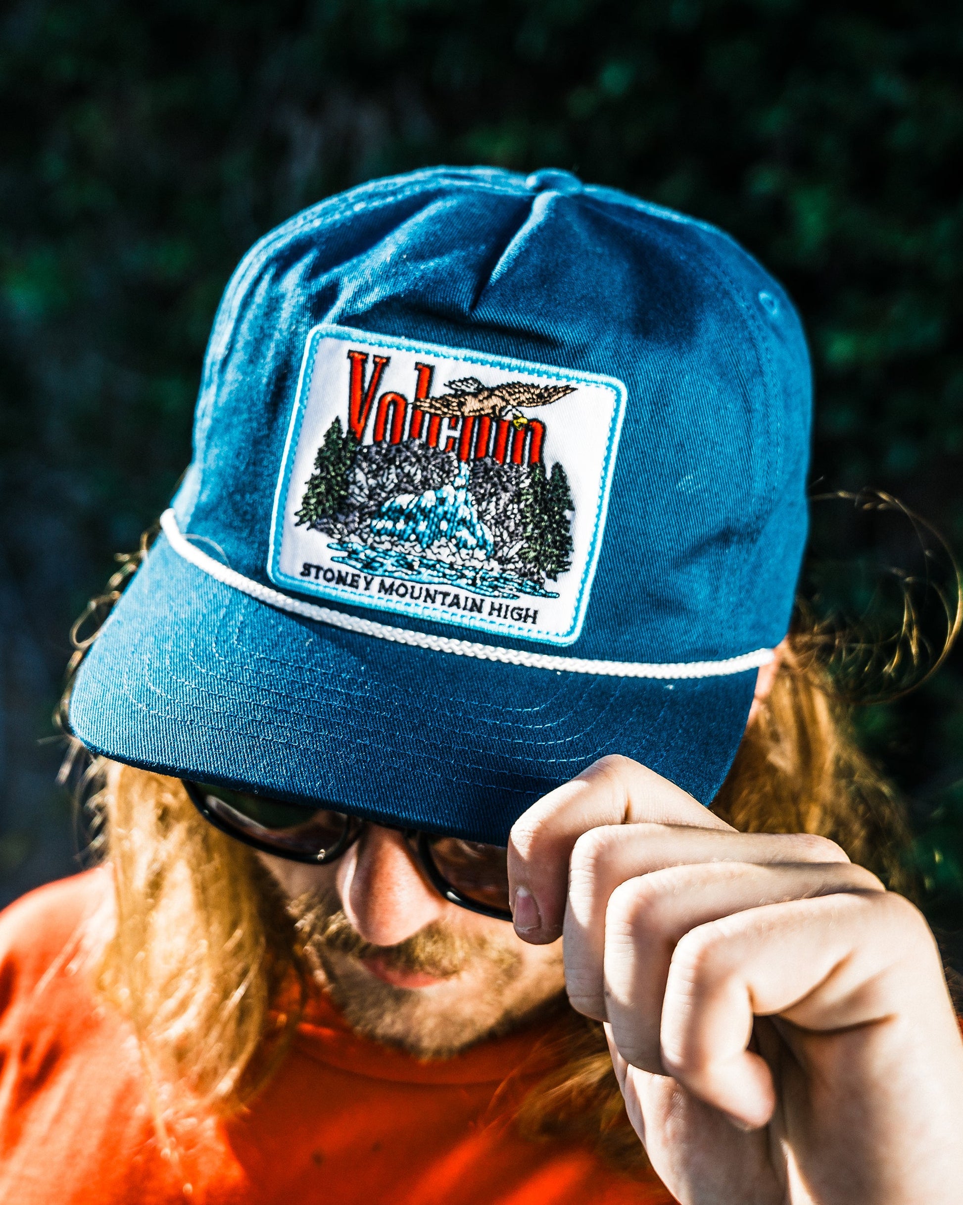 A person with long hair and sunglasses adjusts the brim of the Volcom Men's Stoney Mountain Hi Adjustable Hat, featuring an aqua blue cotton twill build and a branded patch, set against a dark, blurred background.